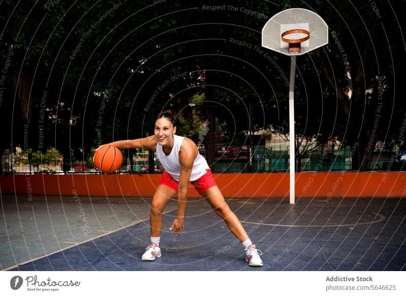 Ganzer Körper einer sportlichen, fröhlichen jungen Frau in Sportkleidung, die wegschaut, während sie an einem sonnigen Sommertag auf dem Platz Basketball spielt