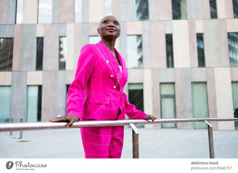 Lächelnde elegante schwarze Frau mit rasiertem Kopf in rosa Outfit steht auf der Straße vor einem Gebäude trendy Aussehen Stil Vorschein verträumt Jacke