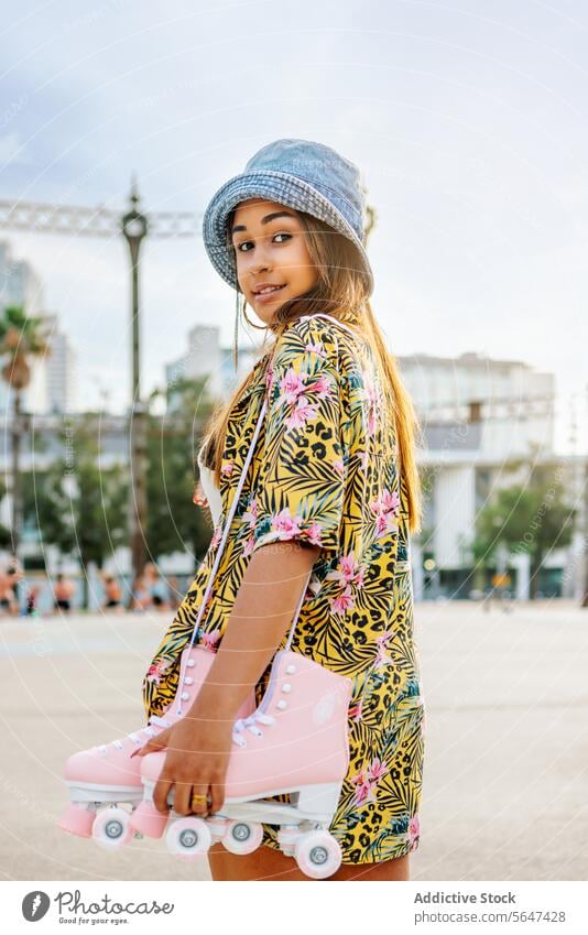 Charmantes Mädchen im Sommeroutfit hält Rollschuhschuhe auf der Straße Frau trendy Skater Mode Hut Stil Großstadt positiv hell selbstbewusst Kleid Stadt modern