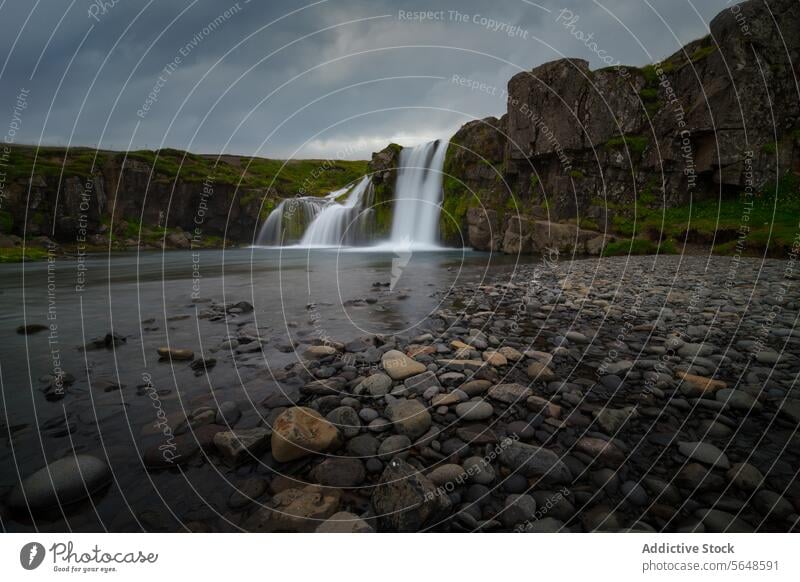 Bedeckter Himmel über einem friedlichen Wasserfall, umgeben von dunklen Klippen und verstreuten Felsen im Vordergrund in Island bedeckt Landschaft Natur