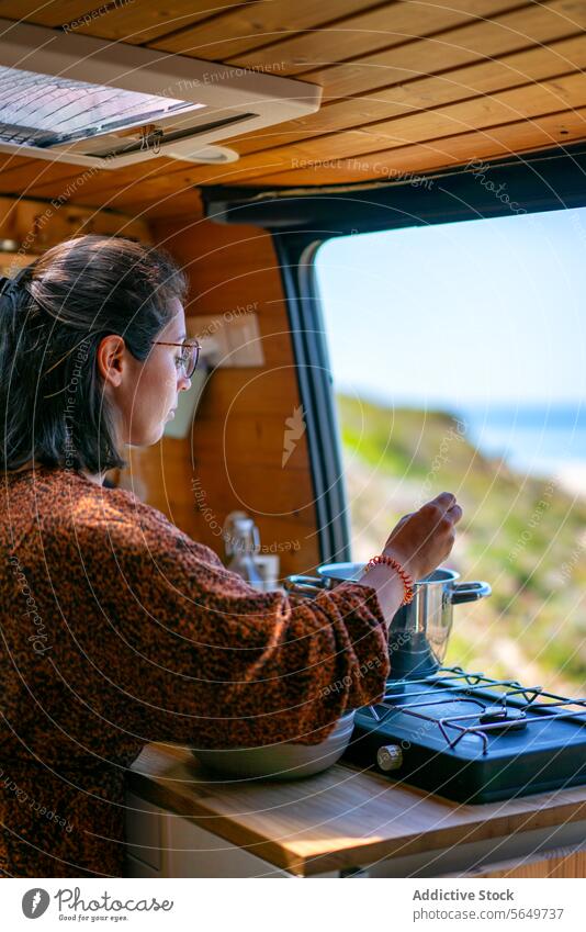 Frau kocht Essen in Wohnwagenküche Reisender Koch Lebensmittel Wohnmobil Kleintransporter Herd Fenster Küche Ausflug Seitenansicht fokussiert Freizeitkleidung