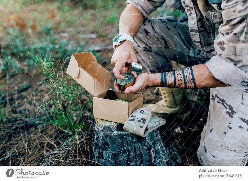 Anonymer Militärsoldat mit Rucksack, der eine Konservendose öffnet, kniend neben einer Kiste auf einem Baum Mann Kommando Besteck Kasten Tarnung Wald Uniform