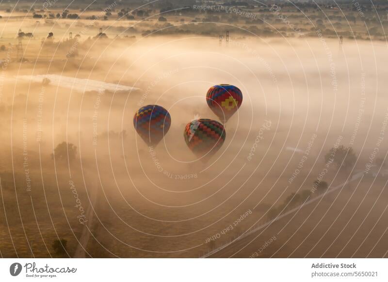 Von oben steigen mehrere bunte Heißluftballons über einer nebelverhangenen Landschaft auf, während die Sonne ein sanftes Licht auf die Szene wirft Ballone Nebel