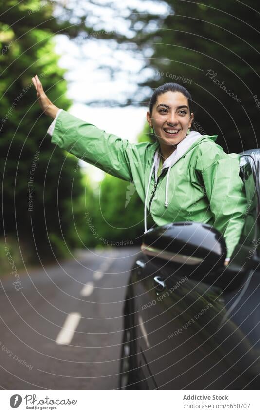 Glückliche junge Frau mit ausgestreckten Armen, die wegschaut und sich aus dem Autofenster lehnt, während sie einen Road Trip im Wald genießt sorgenfrei