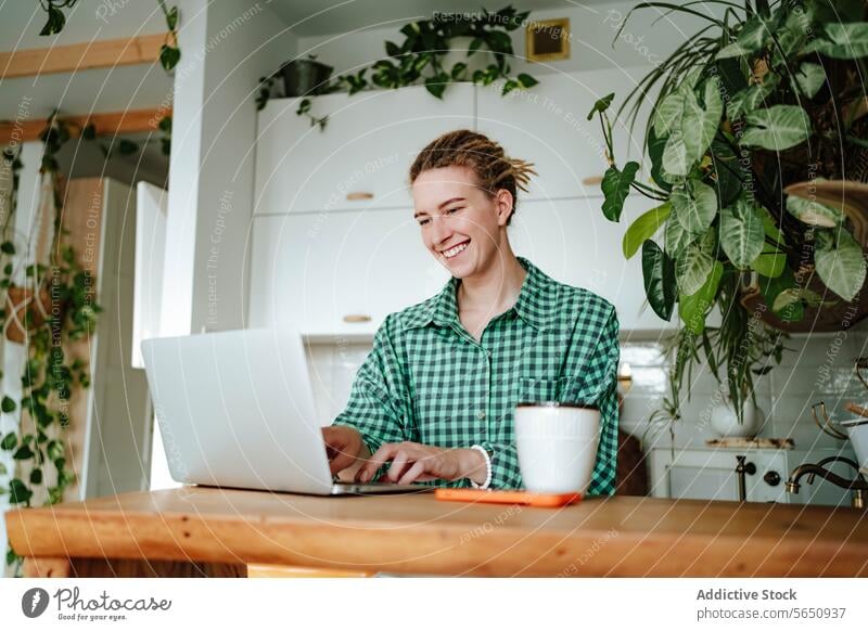 Glückliche junge Frau arbeitet am Laptop am Tisch in der dekorierten Küche zu Hause freiberuflich Arbeit abgelegen Inbetriebnahme online sitzen kariertes Hemd