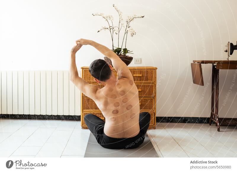 Junger Mann sitzt auf dem Boden und streckt die Hände beim Rehabilitationstraining in die Höhe Dehnung hochreichen Training ohne Hemd heimwärts sitzen Unterlage