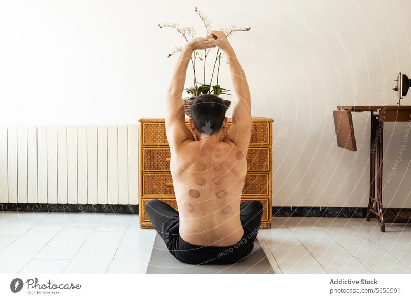 Junger Mann sitzt auf dem Boden und streckt die Hände beim Rehabilitationstraining in die Höhe Dehnung hochreichen Training ohne Hemd heimwärts sitzen Unterlage