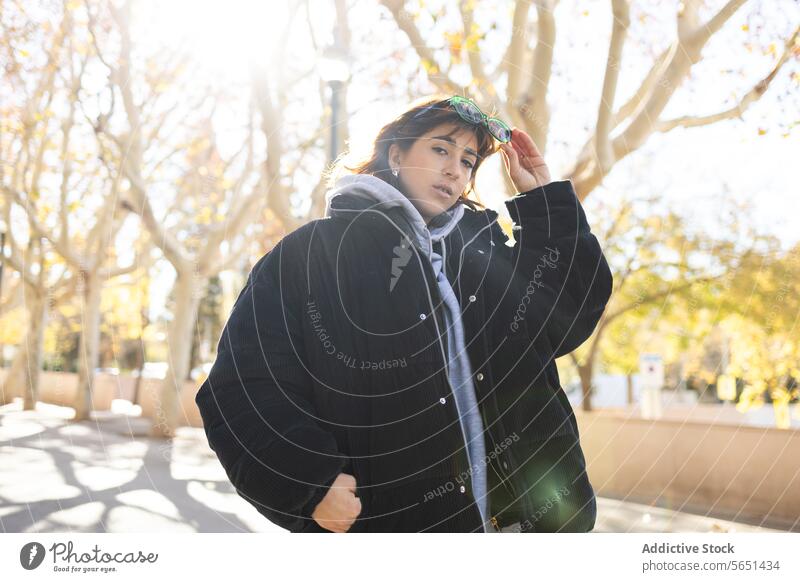 Junge Frau genießt einen sonnigen Herbsttag im Freien jung sonnenbeschienen Park Jacke Sonnenbrille lässig Mode Stil Baum Natur Tageslicht Freizeit entspannt