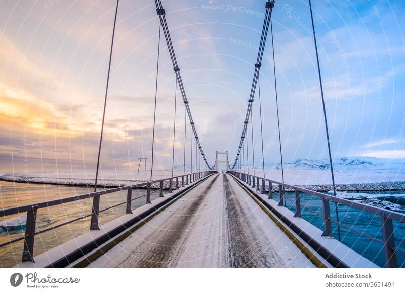 Hängebrücke mit Stahlseilen über einen zugefrorenen Fluss vor der Kulisse schneebedeckter Berge in der Abenddämmerung Kettenbrücke zugefrorener Fluss Schnee