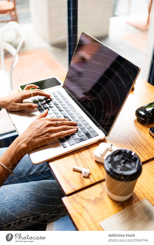 Abgeschnittene, nicht erkennbare Frauenhände, die in einem Café auf einem Laptop tippen, mit einem Smartphone, Kopfhörern und einer Kaffeetasse auf dem Tisch während einer Fernarbeit in Chiang Mai, Thailand