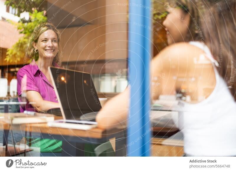 Durch das Fenster genießt eine Frau in einem leuchtend lila Hemd die Fernarbeit an ihrem Laptop in einem Café mit ihrem multiethnischen Kollegen in Chiang Mai, Thailand