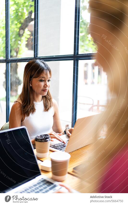 Zwei multiethnische Kollegen konzentrieren sich auf ihre Laptops in einem modernen Café mit großen Fenstern und Blick auf die Stadt, während sie in Chiang Mai, Thailand, per Fernzugriff arbeiten