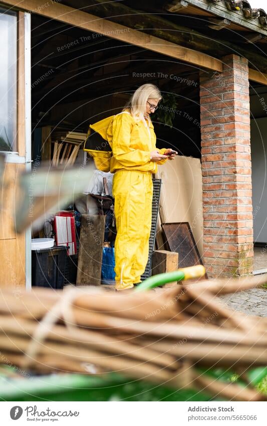 Fokussierte Imkerin in gelbem Overall, die ein Mobiltelefon benutzt, während sie im Bienenstock vor einem Gebäude arbeitet Frau Smartphone Bienenkorb gesamt