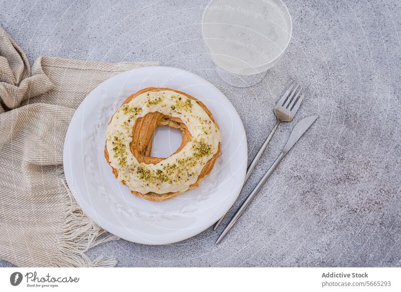 Ringförmiger Brandteig mit weißem Zuckerguss von oben, bestreut mit gehackten Pistazien, präsentiert auf einem weißen Teller mit Gabel und Glas, platziert auf einem beigen Tischset auf einer strukturierten grauen Oberfläche