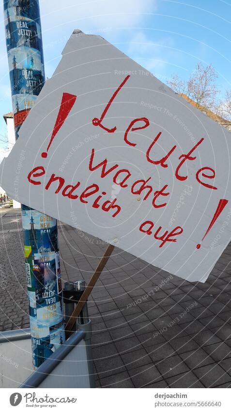 Ein beschriebenes Schild auf einem Holz  Stock,  aufgesteckt an einem Metall Pfahl , mit der Schrift in Rot  : ! Leute wacht endlich auf !