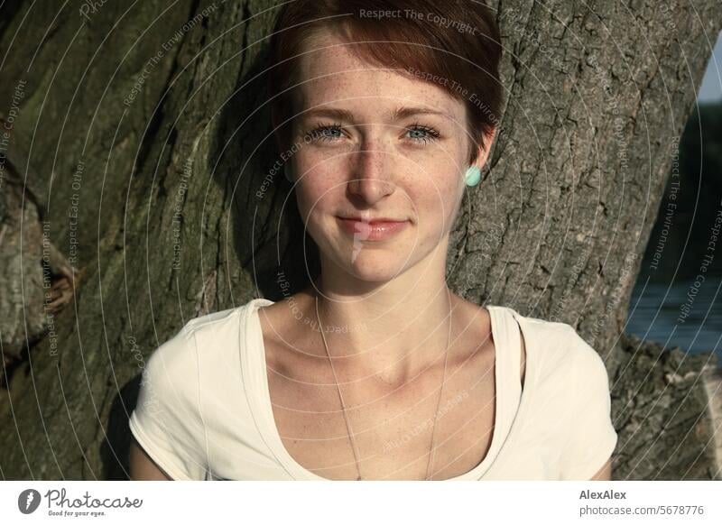 Portrait einer jungen, schlanken, schönen Frau mit Sommersprossen in einem weißem Top, an einem Baum gelehnt aschlank Sommerabend Abend BAum See Lächeln Freude