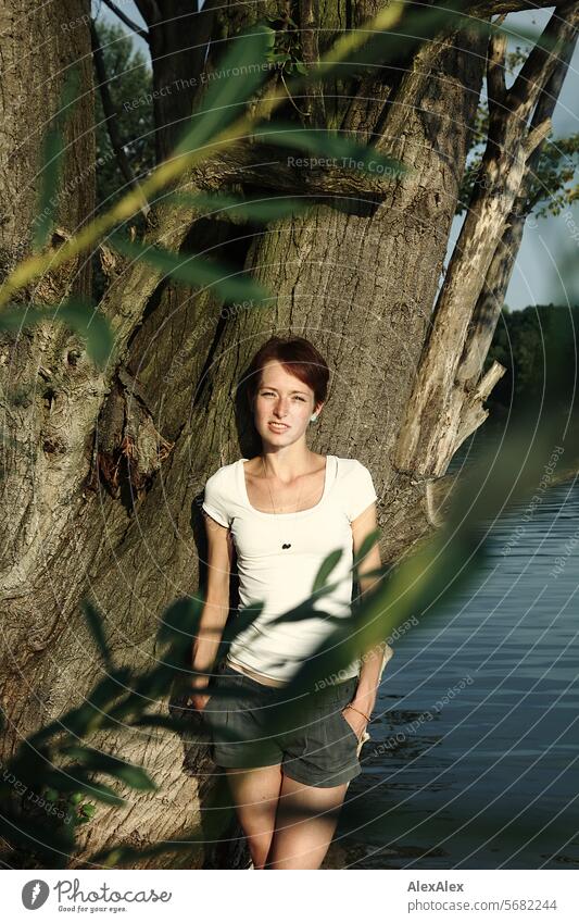 Portrait einer jungen, schlanken, schönen Frau mit Sommersprossen in einem weißem Top und Shorts, an einem Baum gelehnt und leicht verdeckt von einem Zweig mit Blättern