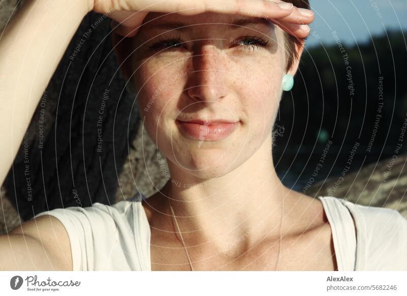 Portrait einer jungen, schlanken, schönen Frau mit Sommersprossen in einem weißem Top, an einem Baum gelehnt und hält sich die Hand über die Augenbrauen, weil die Sonne blendet