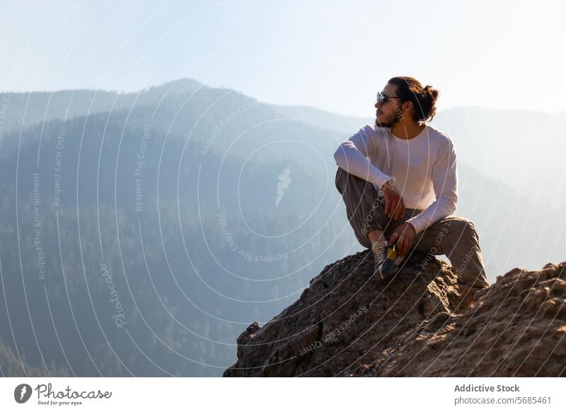 Junger Mann sitzt auf einer Klippe in der Nähe der nebligen Berge sitzen Berge u. Gebirge bewundern Tourist lässig Outfit Vollbart Sonnenbrille felsig Natur