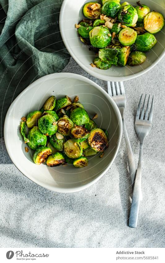 Draufsicht auf zwei Schalen mit gegrilltem, mit Knoblauch und Gewürzen aromatisiertem Rosenkohl, begleitet von silbernen Gabeln auf einem strukturierten Tischtuch