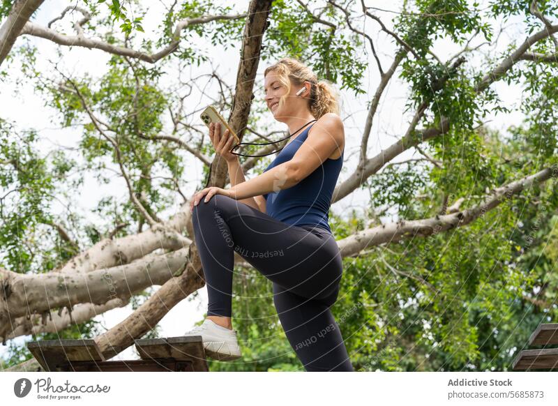 Lächelnde Frau mit Smartphone in der Nähe von Baumzweigen Fitness Training Schiffsplanken schlank Sportkleidung Turnschuh Mobile benutzend Sportlerin Übung