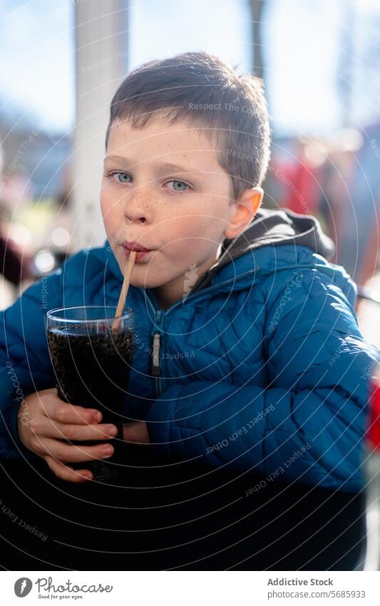 Ein Junge in einer blauen Jacke schaut in die Kamera, während er an einem Tisch eine dunkle Limonade durch einen Strohhalm schlürft Soda trinken schlürfend jung