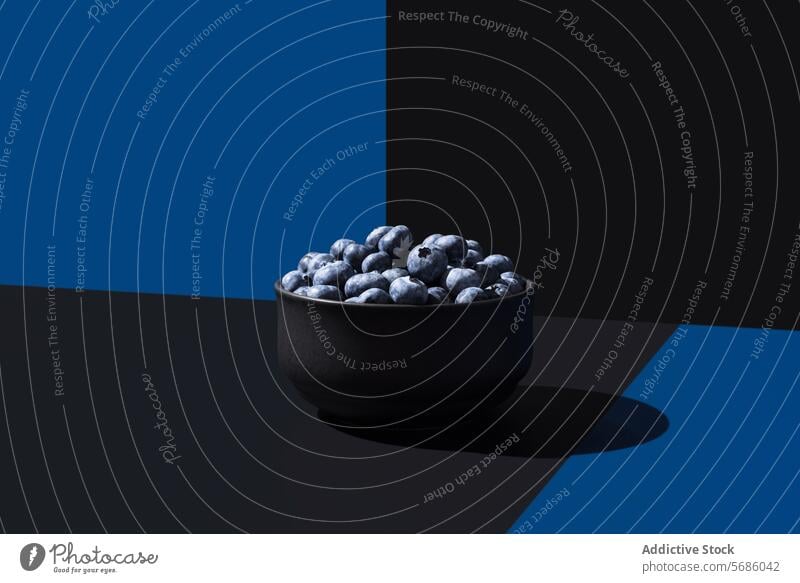 Eine minimalistische Komposition mit einer schwarzen Schale mit Blaubeeren auf einem geteilten blauen und schwarzen Hintergrund mit dramatischer Beleuchtung