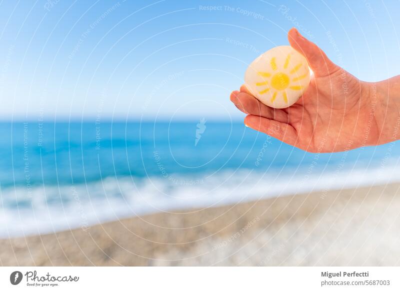 Kinderhand, die einen Strandstein mit einer darauf gezeichneten Sonne hält, ein unscharfer Strand im Hintergrund. Hand Stein Sand Urlaub Wasser Sommer Schönheit