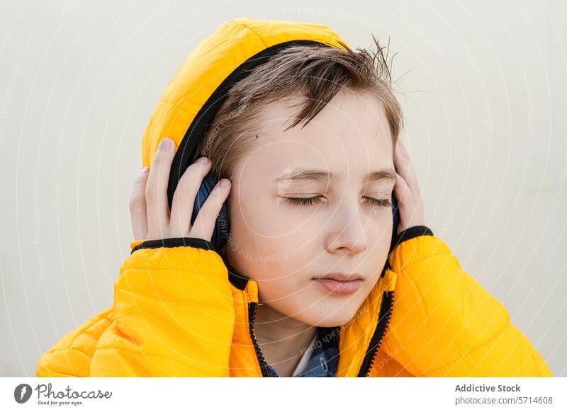 Ein nachdenklicher Junge in einer auffälligen gelben Jacke genießt Musik über seine Kopfhörer vor einem weichen, neutralen Hintergrund hören beschaulich