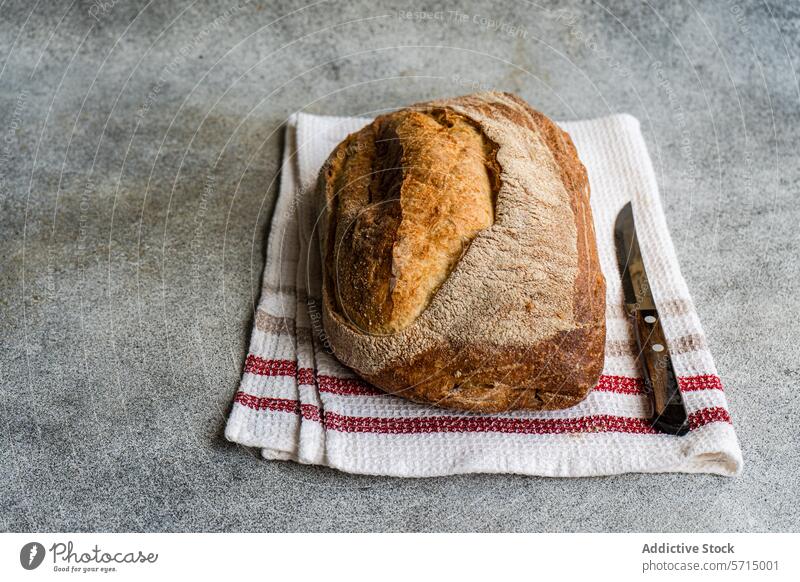 Draufsicht auf goldbraunes Roggensauerteigbrot auf einem weißen Tuch mit roten Streifen, daneben ein Brotmesser auf einer strukturierten Oberfläche Sauerteig