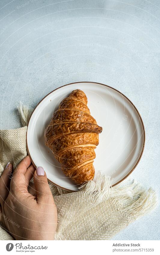 Draufsicht auf eine anonyme Person, die einen Teller mit einem frisch gebackenen Croissant auf einem strukturierten Tuch in der Hand hält und damit einen einfachen und gemütlichen Frühstücksmoment festhält