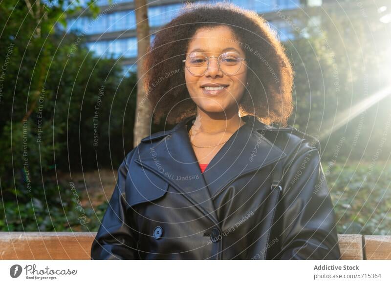 Strahlende junge Frau mit Brille, die auf einer Parkbank lächelt, während das Sonnenlicht durch die Bäume und ein Gebäude im Hintergrund fällt Lächeln im Freien