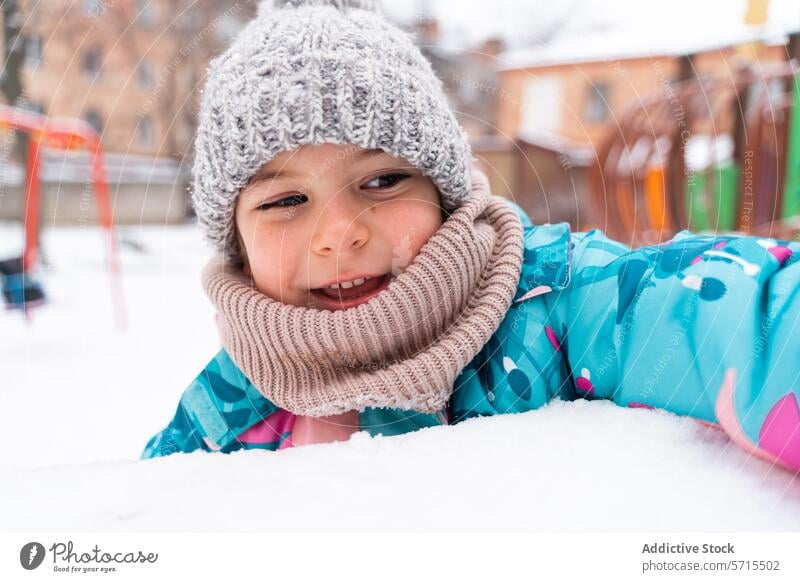 Lächelndes Kind im Schnee liegend, mit Strickmütze und bunter Jacke, mit Blick auf den Spielplatz Winter bunte Jacke im Freien kalt Glück Spaß Schal Freude