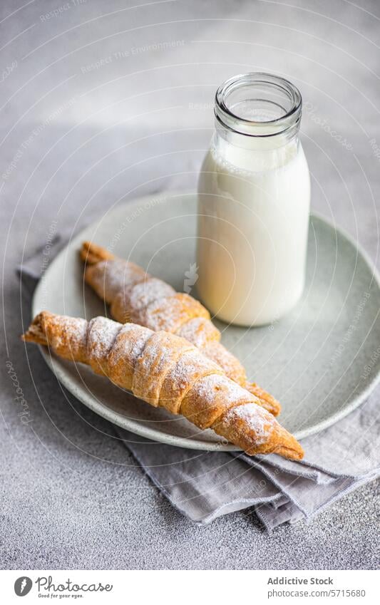 Zwei mit hausgemachter Marmelade gefüllte und mit Puderzucker bestäubte Törtchen, dazu eine Flasche Milch, auf einem Keramikteller über einer grauen Serviette und einer Arbeitsplatte