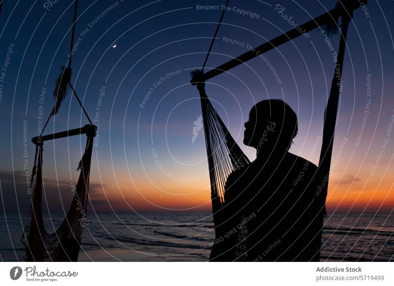 Anonyme Person Silhouette steht neben einer Hängematte am Strand, mit dem letzten Licht des Sonnenuntergangs und eine Mondsichel über Dämmerung Gelassenheit