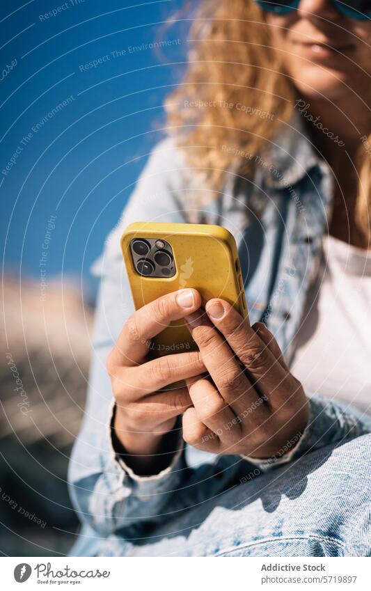 Eine Nahaufnahme zeigt eine Frau mit Sonnenbrille, die ein Smartphone mit einem gelben Gehäuse in der Hand hält, wobei der Fokus auf den Händen liegt, die mit dem Gerät unter einem klaren blauen Himmel interagieren.