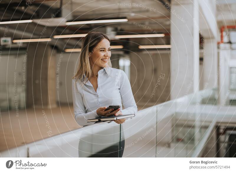 Junge Frau mit Notebook, die ein Mobiltelefon benutzt und in einem modernen Büroflur steht Erwachsener attraktiv schön Business Geschäftsfrau lässig Unternehmen