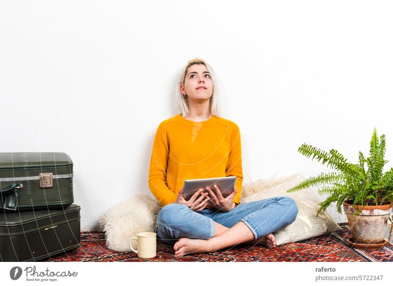 Nachdenkliche Frau mit Tablet und Blick nach oben zu Hause. Tablette Kontemplation Inspiration gelber Pullover heimwärts Denken besinnlich neugierig Denim-Jeans