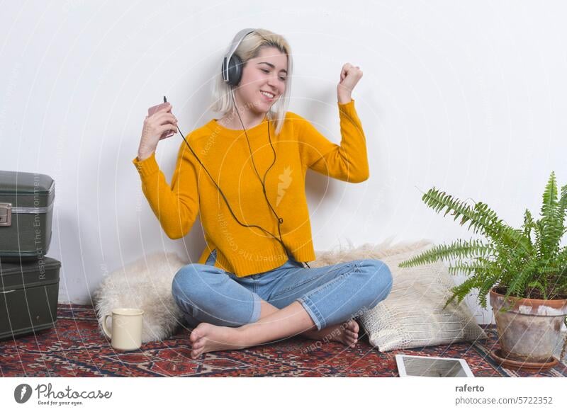 Frau mit Kopfhörern tanzt und hält Telefon. Tanzen Smartphone Glück gelber Pullover Musik Freude heimwärts Energie lässig Denim-Jeans Sitzen Vorleger Genuss