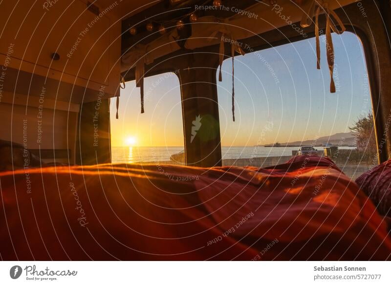 Blick aus einem selbstgebauten Wohnmobil am Strand des Mittelmeers mit farbenfrohem Sonnenuntergang über der Wasseroberfläche, Andalusien, Spanien