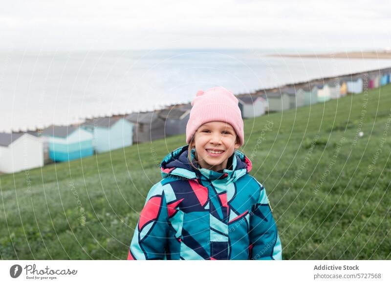 Ein fröhliches junges Mädchen mit einer rosa Mütze und einem bunten Mantel lächelt warm auf einem grünen Hügel mit Blick auf die kultigen Strandhütten von Whitstable, UK