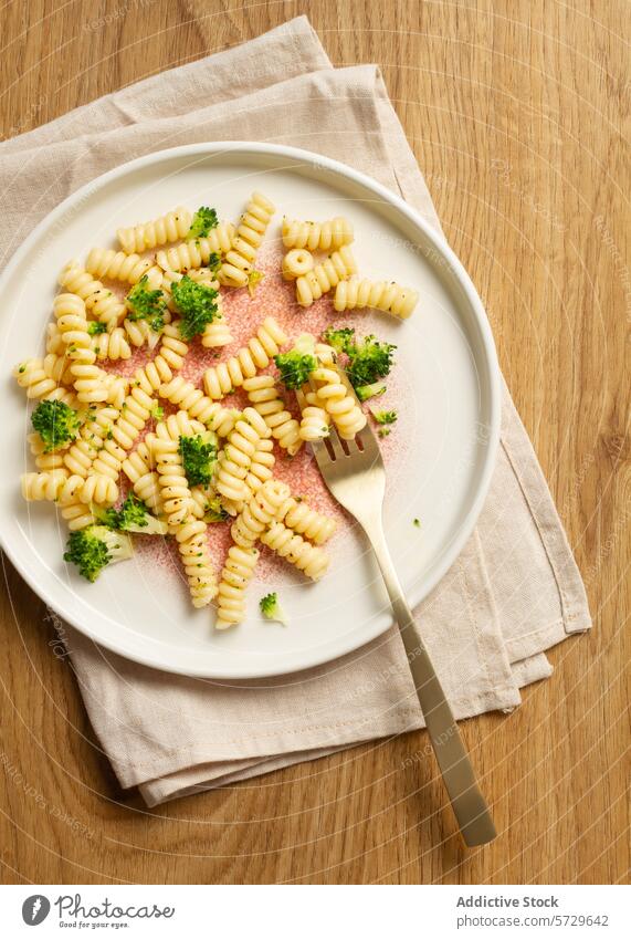 Eleganter gesprenkelter Teller mit einer Portion Fusilli-Nudeln mit Brokkoliröschen, gepaart mit einer goldenen Gabel auf einer beigen Serviette und einem hölzernen Hintergrund