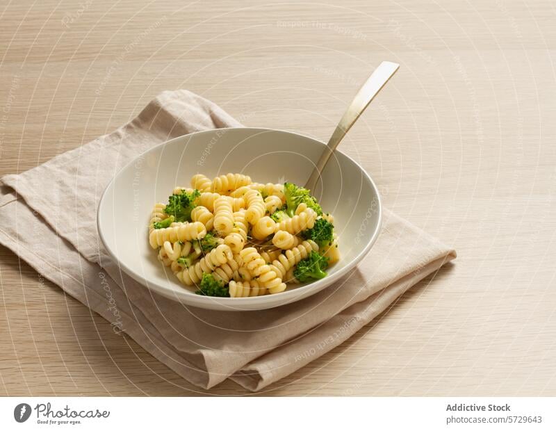 Eine Schale mit Fusilli-Nudeln und gedünstetem Brokkoli, dazu eine beigefarbene Leinenserviette und eine moderne Gabel auf einer Holzfläche Spiralnudel Spätzle