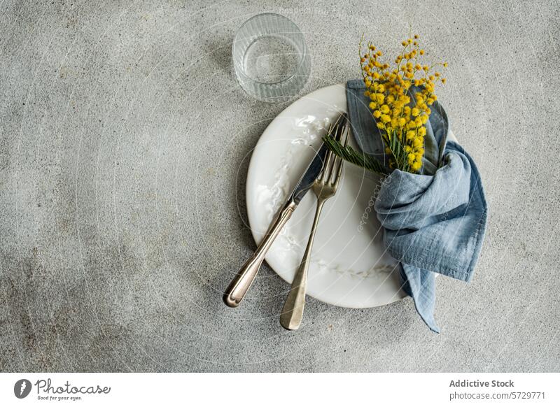 Elegante Tischdekoration mit einem weißen, verzierten Teller, altem Silberbesteck, einer blauen Leinenserviette und einem leuchtend gelben Mimosenstrauß auf einem strukturierten Hintergrund