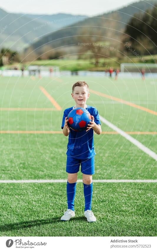 Junger Fußballspieler hält einen Ball auf dem Feld Spieler Uniform Sport Platz Jugend Kind sportlich aktiv im Freien Beteiligung Lächeln Selbstvertrauen Gras