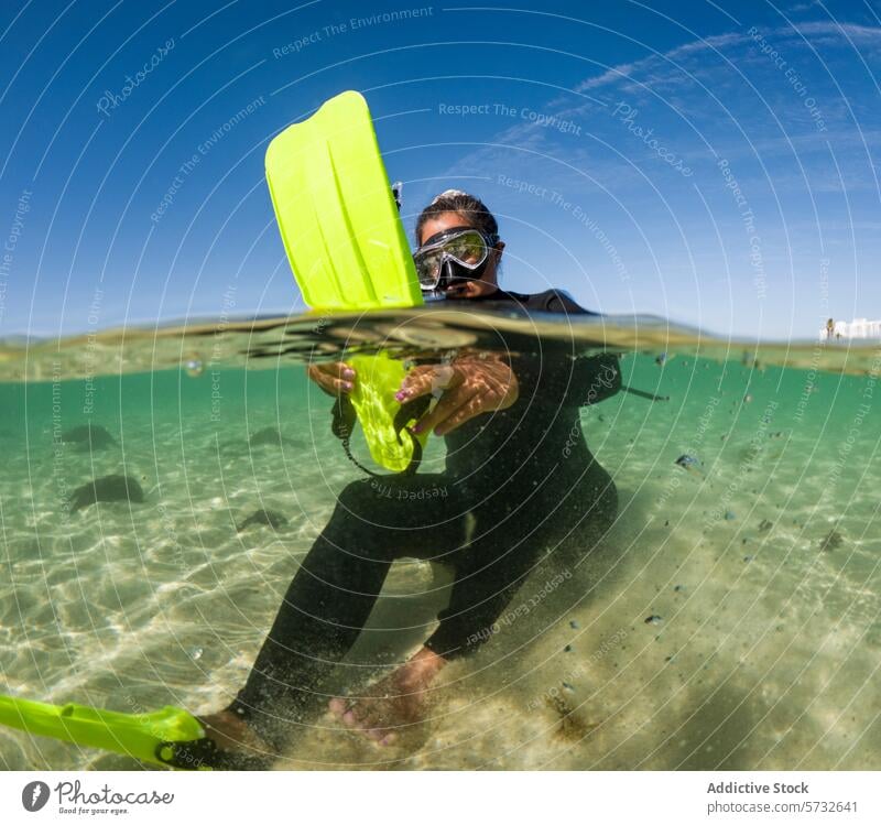 Ein Schnorchler in einem schwarzen Anzug mit einer leuchtend gelben Flosse schaut durch eine Tauchermaske, halb untergetaucht im sonnenbeschienenen, klaren Meerwasser