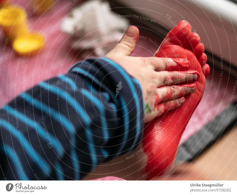 Ein Kind malt mit seiner Hand und roter Fingerfarbe seinen Fuß an malen Kunst Kinderkunst Leben mit Kindern Farbfoto Innenaufnahme am Fenster