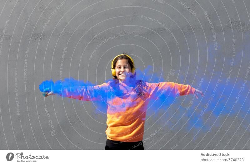 Fröhliches Mädchen spielt mit buntem Pulver an einem sonnigen Tag freudig farbenfroh blau orange Kopfhörer Lächeln Spielen pulsierend grau Hintergrund