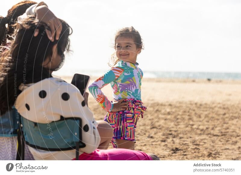 Ein fröhliches junges Mädchen posiert mit den Händen auf den Hüften und strahlt vor Zuversicht an einem sonnigen Strandtag, während ihre Familie ein Foto von ihr macht