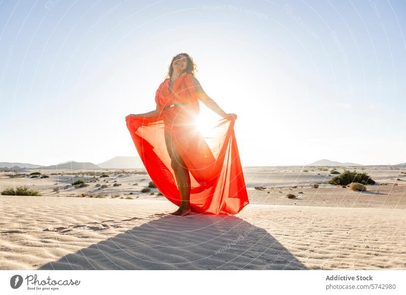 Frau in leuchtend rotem Kleid steht auf sandigen Dünen wüst Dunes Sand elegant Pose Sonnenlicht golden fließend Gewebe pulsierend Mode im Freien Gelassenheit